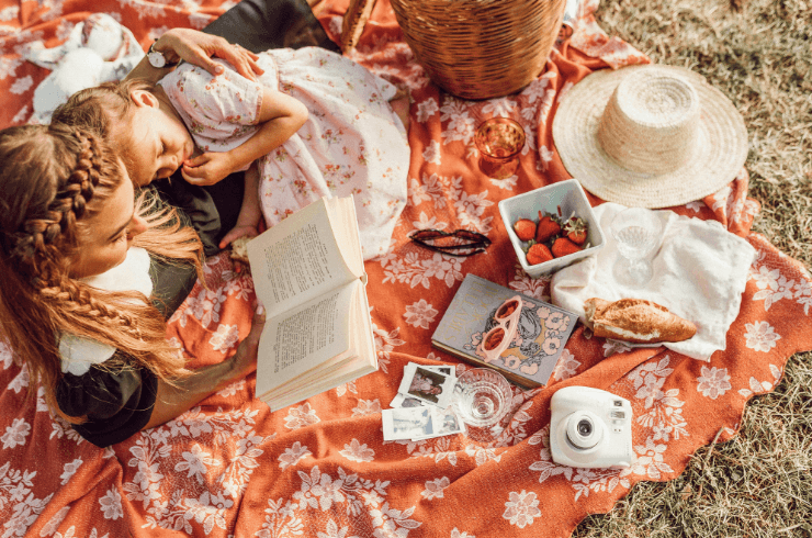 A girl and her mother reading a book on a picnic blanket with fruits and a croissant visible