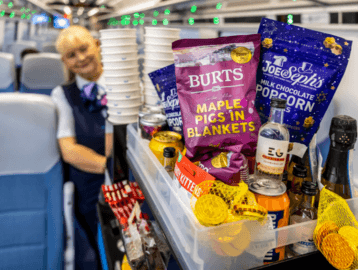 A TransPennine Express staff member pushing a food trolley where Burt's Maple Pigs in Blankets and Joe & Sephs milk chocolate popcorn can be seen.